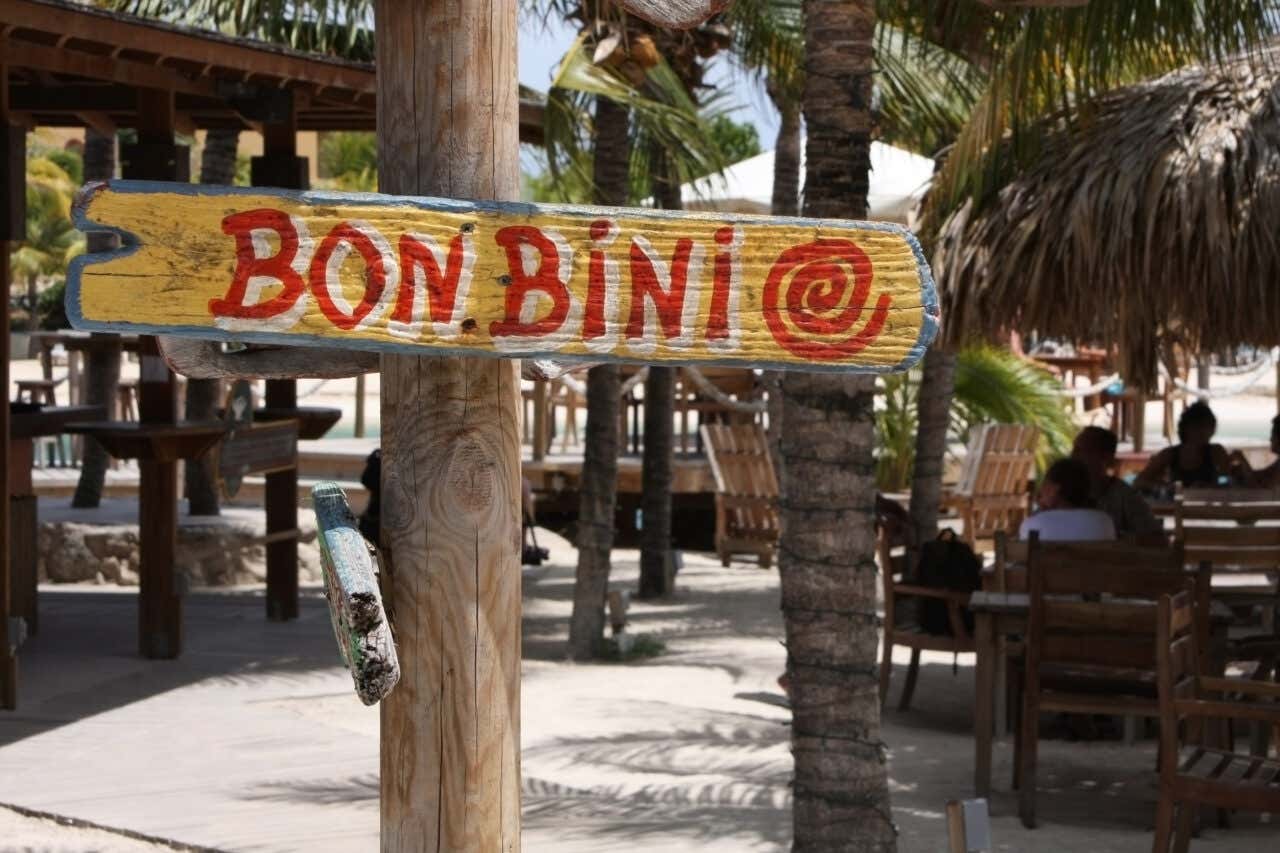 A sign reading Bonbini with a mixture of capitalised and uncapitalised letters, written in red on a wooden sign, pointing to the viewers right. In the background one can see various trees and some  dining tables on a beach.