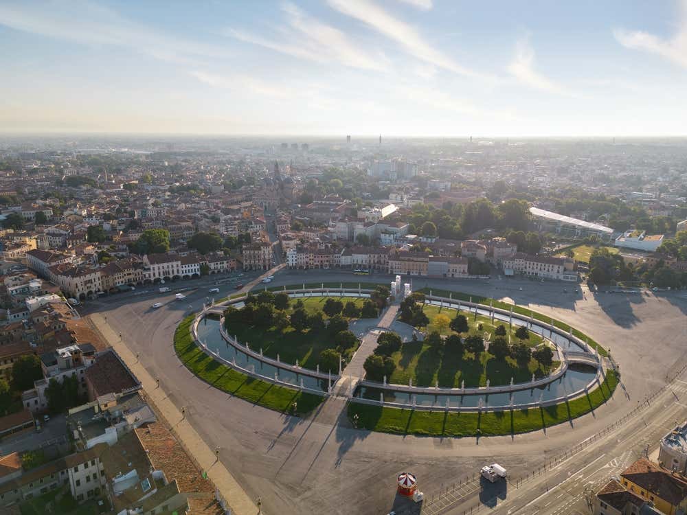 Veduta aerea dell’ampio Prato della Valle a Padova: una grande piazza ellittica con un canale che la circonda, ponti che collegano l’isola centrale (Isola Memmia) al perimetro esterno, e statue allineate lungo il bordo del canale, mentre sullo sfondo si intravede il tessuto urbano della città