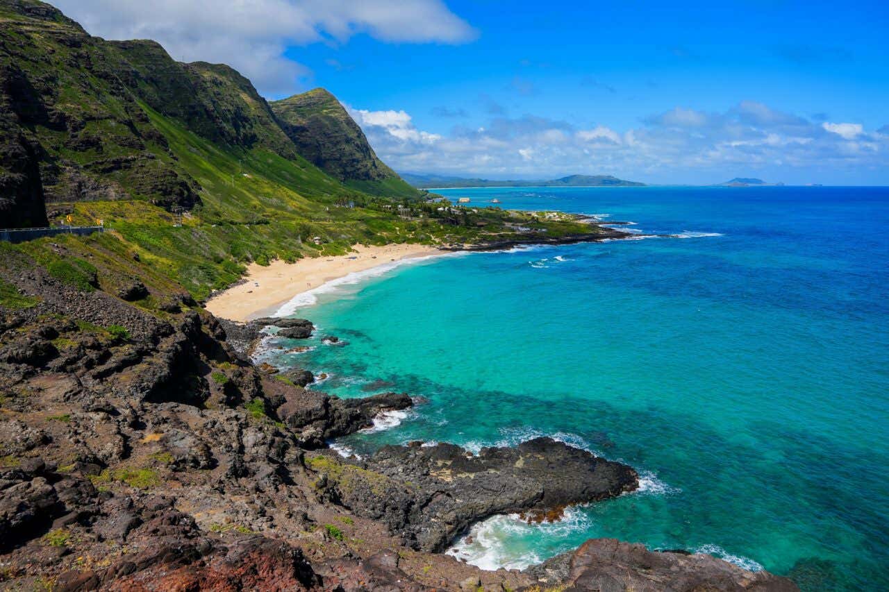 A high-angle, scenic view of a beautiful, remote tropical beach cove. The bright turquoise ocean water meets a narrow strip of golden sand, backed by steep, lush green mountains under a bright blue, partially cloudy sky.