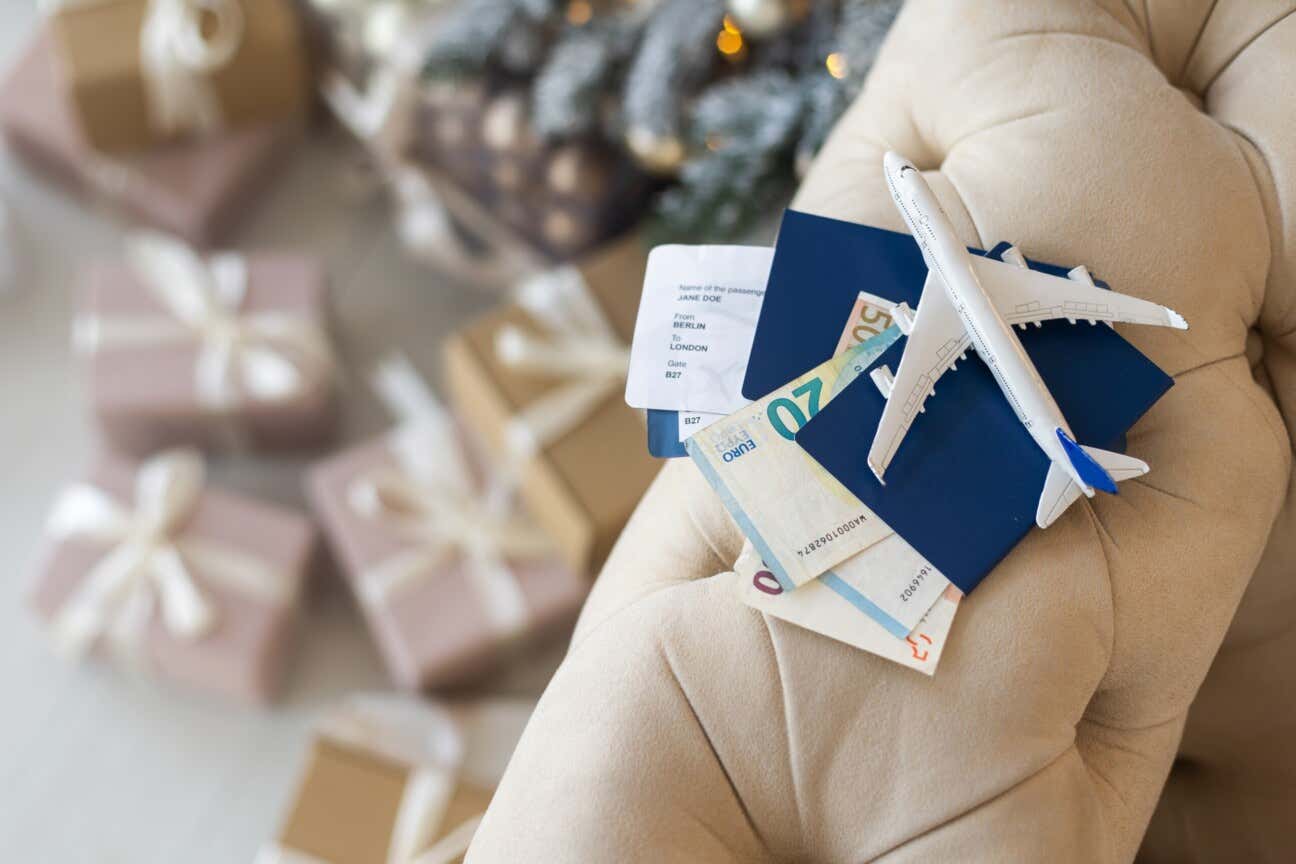 Un piccolo aeroplanino di plastica poggiato sul bracciolo di un divano insieme ai biglietti di un volo e contanti, sullo sfondo, ai piedi del divano, regali di Natale e un albero decorato per le Feste