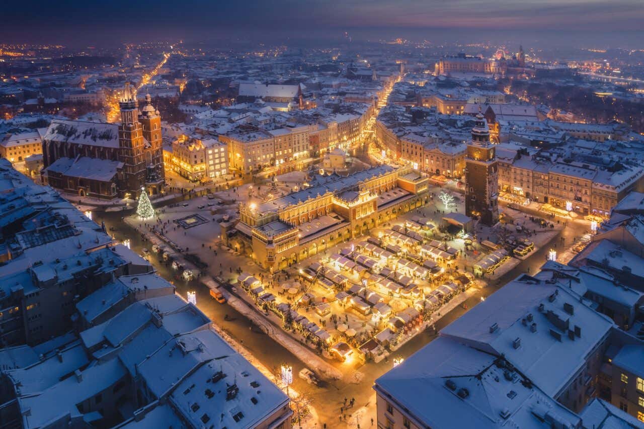 La piazza centrale di Cracovia fotografata dall'alto in una serata d'inverno. Le tetti delle case sono innevati e al centro c'è un mercatino natalizio illuminato da numerose luminarie