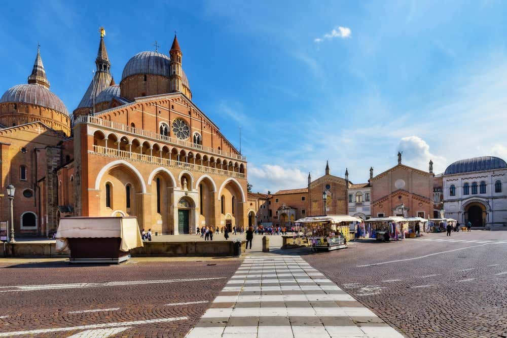 Vista della grande facciata di una chiesa romanica con cupole in ottone, al centro di una piazza di Padova. Di fronte all'ingresso è allestito un piccolo mercatino visitato da turisti e fedeli