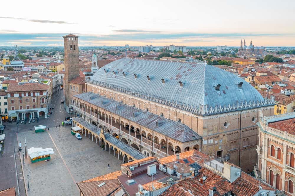 Veduta dall’alto del Palazzo della Ragione a Padova sul lato di Piazza della Frutta: si nota il grande edificio medievale con la sua lunga facciata, le logge ad archi su due piani, e la torre adiacente; sotto, un grande spiazzo ancora vuoto, con alcune bancarelle in allestimento dato che la foto è stata scattata all'alba