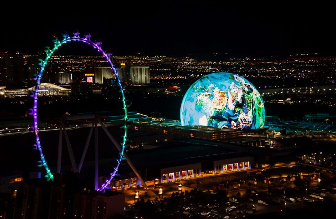 An aerial night view of the Las Vegas Strip, dominated by the massive, glowing spherical structure (The Sphere) displaying a colorful image of Earth, alongside the brightly lit, neon-rainbow High Roller Ferris wheel.