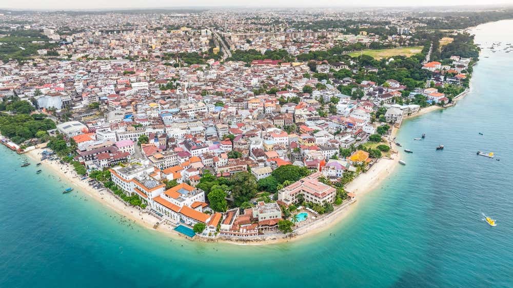Vista della città portuale di Stone Town dall'alto, con palazzi in stile coloniale bassi lungo la riva e barche in mare davanti a ampie spiagge di sabbia bianca