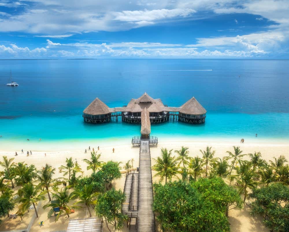 un bungalow di lusso in legno e paglia costruito su palafitte a pochi metri da una spiaggia a Zanzibar, sospeso su un mare turchese e con persone che passeggiano sulla spiaggia