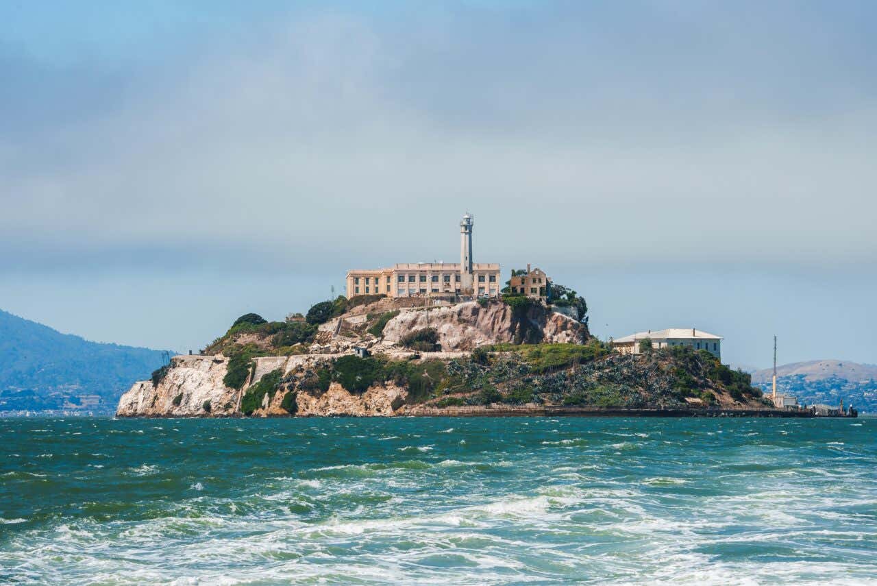 A daytime, long-distance view of Alcatraz Island, showing the former pale-yellow prison building and a white lighthouse atop a rocky, scrub-covered outcrop in the choppy blue waters of San Francisco Bay.