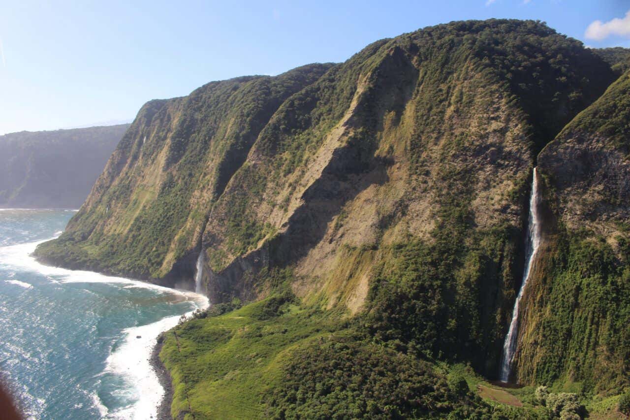 An aerial view of massive, sheer green sea cliffs on the coast of a tropical island. Two dramatic waterfalls cascade hundreds of feet down the steep, lush rock face into the churning blue ocean waves below.