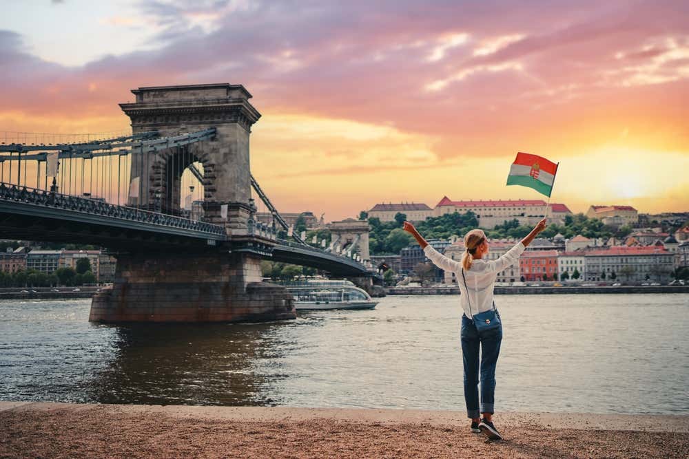 Una ragazza di spalle sulle rive del fiume Danubio, mentre tiene in mano la bandiera di Budapest, durante il tramonto.