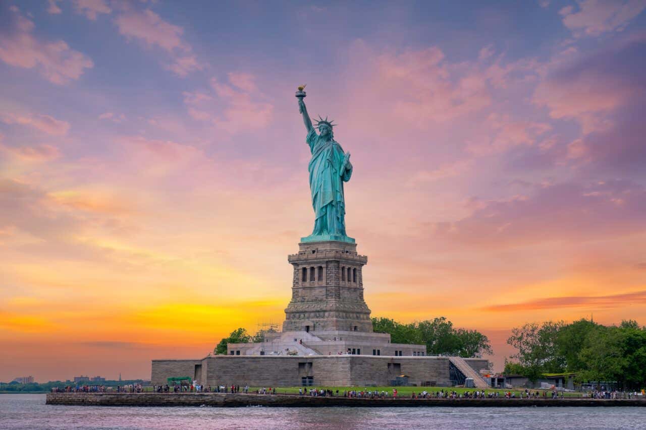 A full view of the Statue of Liberty standing on its pedestal against a dramatic sunset sky with hues of orange, yellow, and purple. The statue is centered and framed by the colorful sky and dark foreground landscaping.