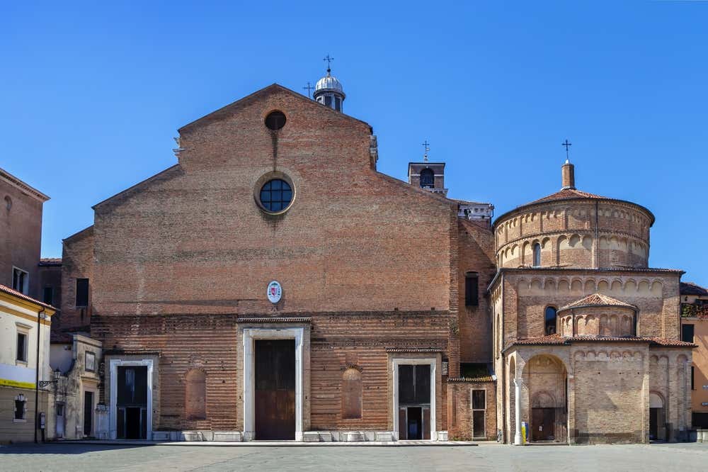 Vista frontale della Cattedrale di Santa Maria Assunta a Padova affiancata dal Battistero: la facciata in mattoni chiari della cattedrale, con ingresso centrale e finestra rotonda, è contornata dalla cupola e dal tamburo del battistero romanico, caratterizzato da archetti e una copertura emisferica. Sullo sfondo, il cielo è terso e luminoso