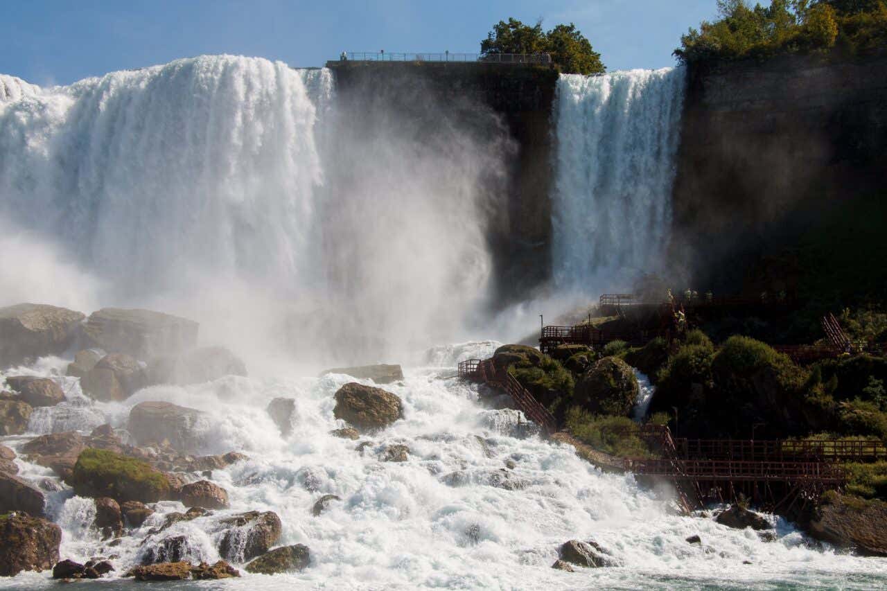 A powerful daytime view of a wide section of a large waterfall (American Falls), showing a massive curtain of white water plunging over a rocky edge into a turbulent, misty pool below, with wooden walkways visible on the right.