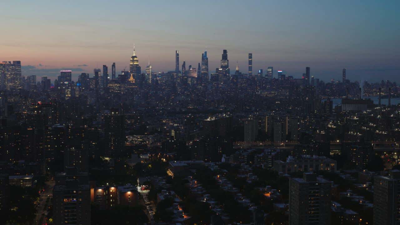 A panoramic view of the New York City skyline at twilight. Many prominent skyscrapers, including the Empire State Building and others in Midtown and Lower Manhattan, rise above the dark city below, illuminated by streetlights and the windows of buildings.