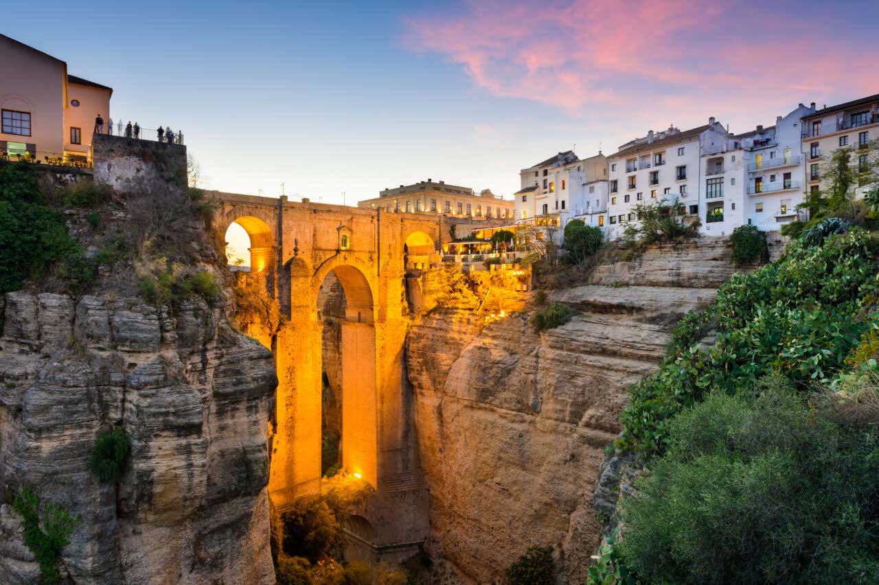 Views of an illuminated stone bridge crossing over a gorge with buildings on both sides at sunset