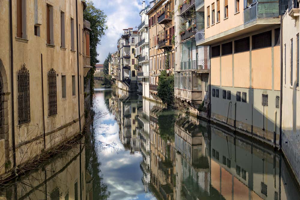 Canale tranquillo nel centro storico di Padova: acqua liscia che riflette le facciate storiche di case in pietra levigata, con finestre classiche e balconcini; le rive sono fiancheggiate da vegetazione verde e un ponte pedonale in pietra collega i due lati della città, creando un’atmosfera romantica e intima