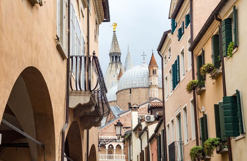 Vicolo stretto nel centro storico di Padova, fiancheggiato da edifici in pietra e mattoni con balconi e finestre su più piani, che si apre in fondo su una vista sulle cupole della Basilica di Sant’Antonio