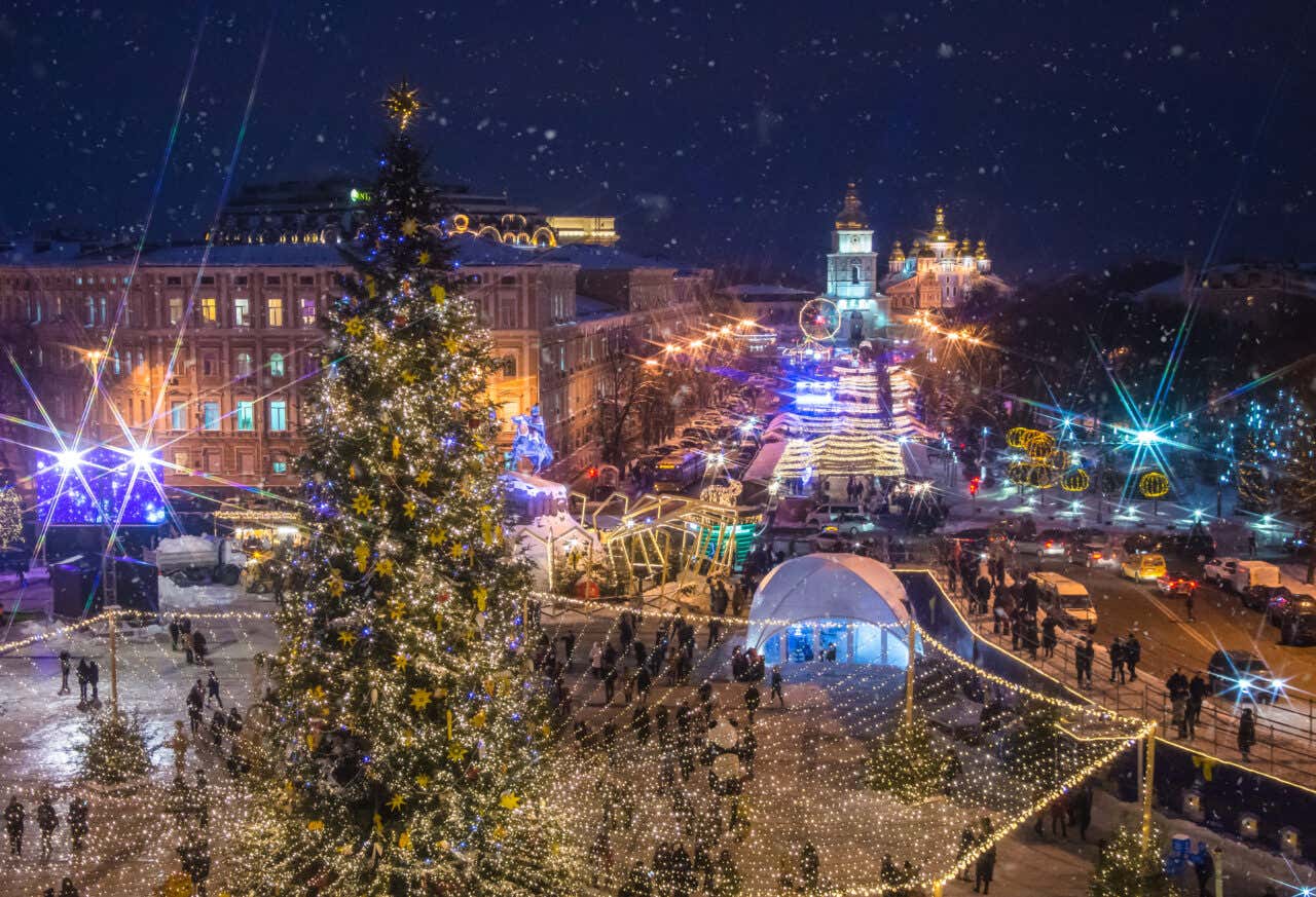 Vista notturna del mercatino di Natale di Sofia con albero illuminato in primo piano, bancarelle e luminarie colorate, Cattedrale di San Alessandro Nevskij sullo sfondo durante una nevicata