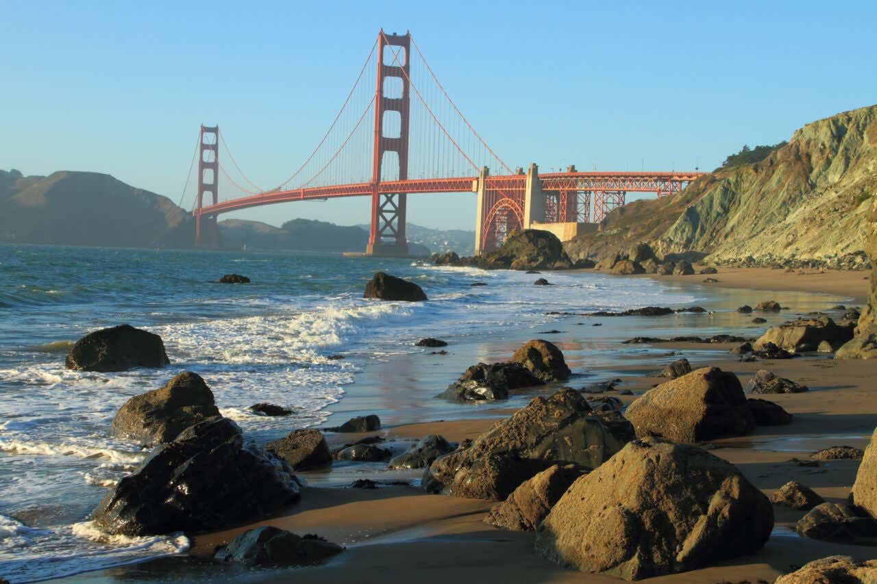 The Golden Gate Bridge seen from a rocky beach at dusk or night, with ocean waves crashing onto the foreground of dark boulders and sand.