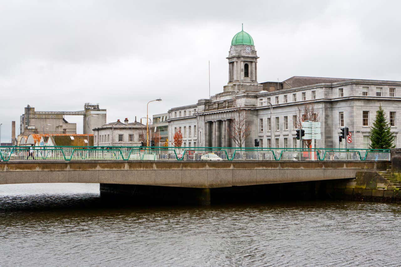 A view of Cork City Hall under a cloudy sky with a bridge seen over the river.