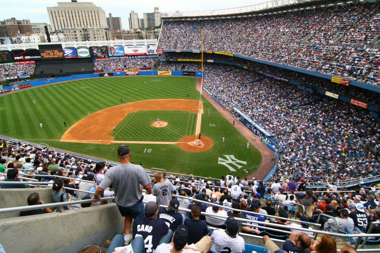 A wide, elevated view of Yankee Stadium during a daytime baseball game. The stadium is packed with fans, with many seated in the foreground. The baseball field is visible, showing a green, striped outfield and the brown infield dirt.