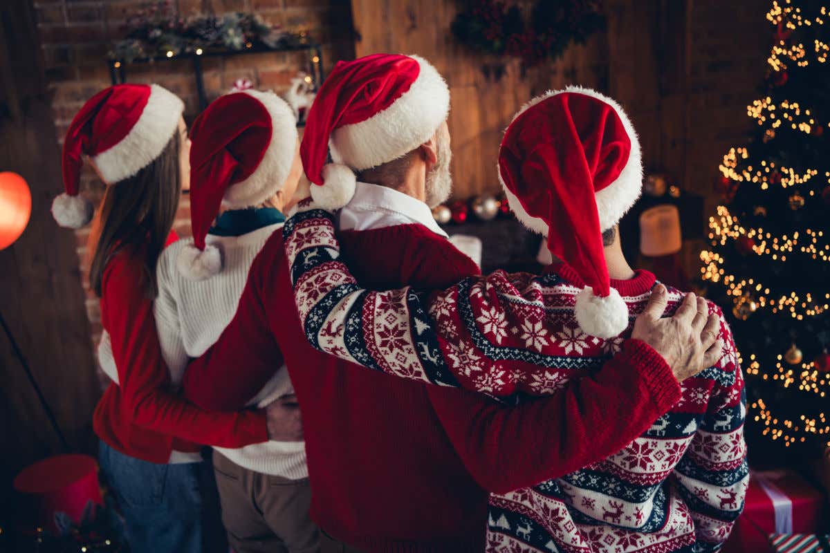 Una familia abrazándose y entrelazados con gorros de Santa Claus en sus cabezas y contemplando un árbol de Navidad en el interior de una casa