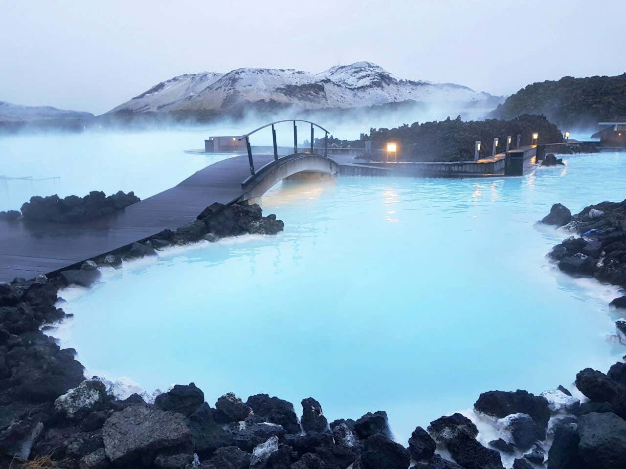 The Blue Lagoon in Iceland on an overcast day with a bridge in the foreground and mountains in the background.