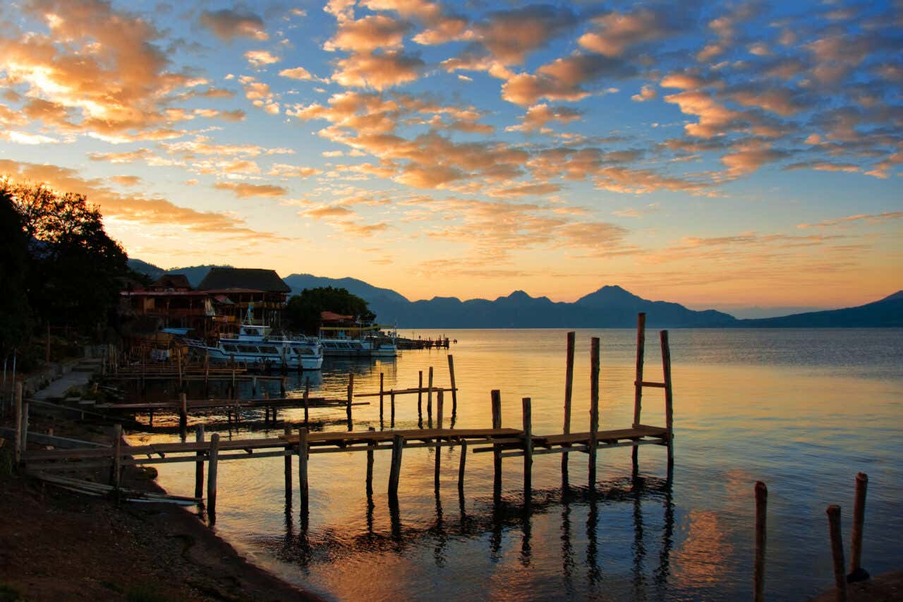 Sunset over a lake, likely Lake Atitlán in Guatemala. The water reflects the warm colors of the sky, which is filled with orange and pink clouds. Wooden docks extend into the water in the foreground, with buildings and mountains silhouetted in the distance.