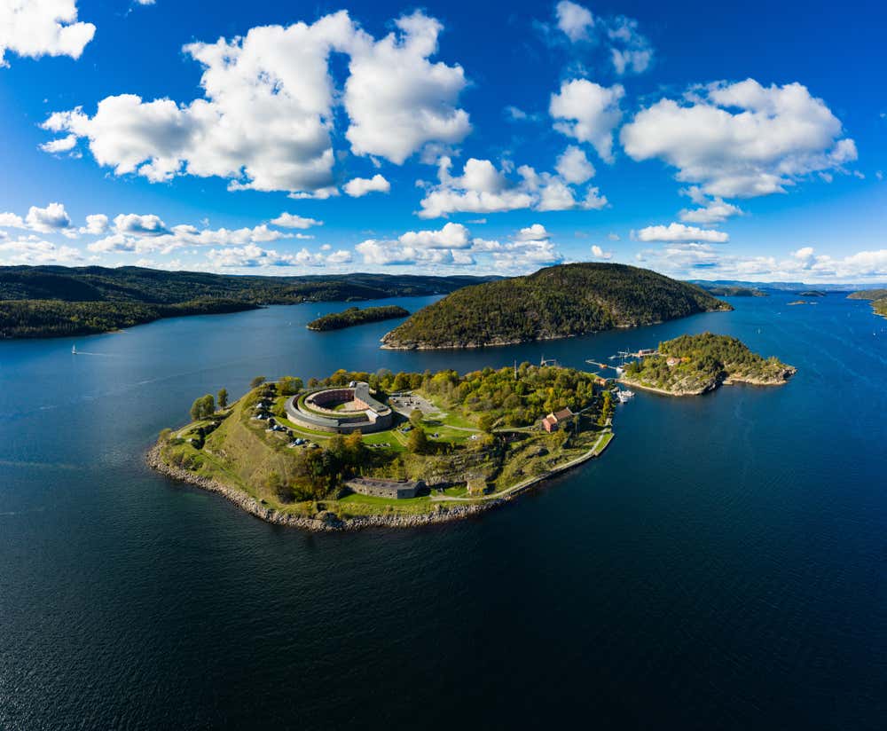 Vista aerea dell'isola di Oscarsborg, nella sua posizione strategica al centro dell'Oslofjorden, in Norvegia. In primo piano, la celebre fortezza circolare ottocentesca immersa nel verde, affiancata da edifici storici in mattoni rossi. Sullo sfondo, le isole boscose e le acque azzurre del fiordo si estendono verso l'orizzonte sotto un cielo estivo con nuvole bianche