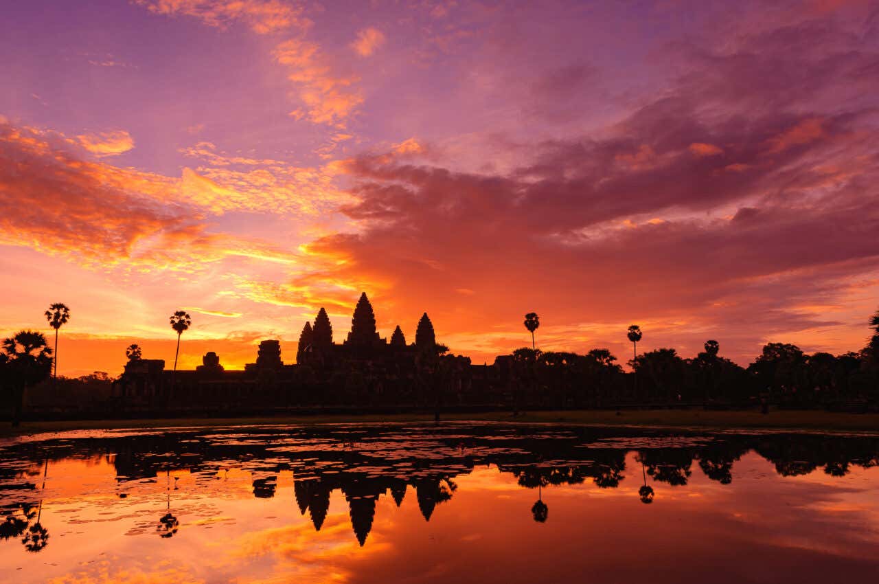 Silhouette of Angkor Wat temple in Cambodia at sunrise or sunset. The temple's towers are reflected in a pool of water in the foreground. The sky is a dramatic mix of fiery orange, magenta, and purple.