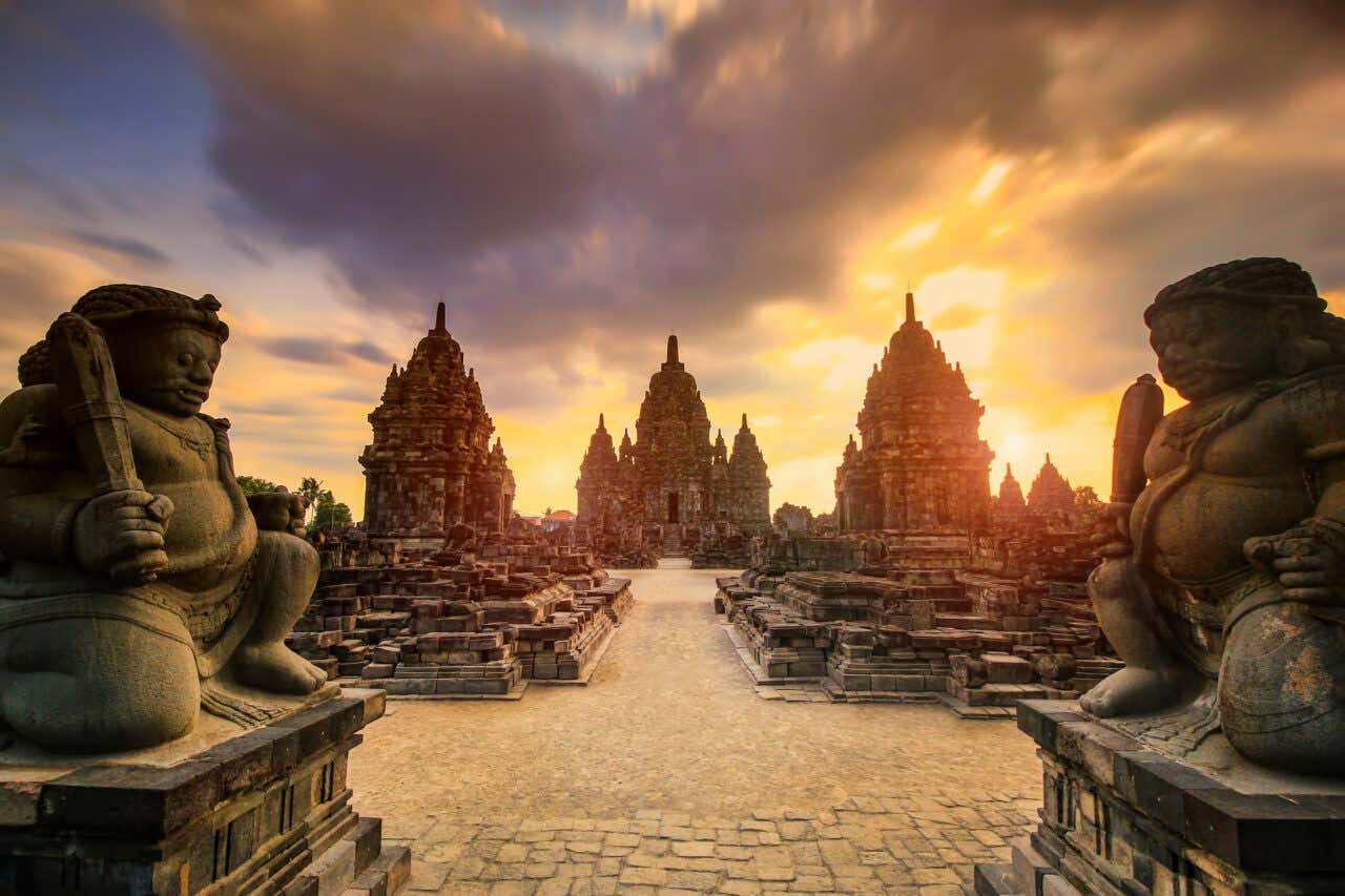 Sunrise or sunset over Candi Sewu, a 9th-century Buddhist temple complex in Java, Indonesia. Two large stone guardian statues stand in the foreground, facing a central path that leads to several richly decorated candi (temple structures) under a dramatic, clouded sky.