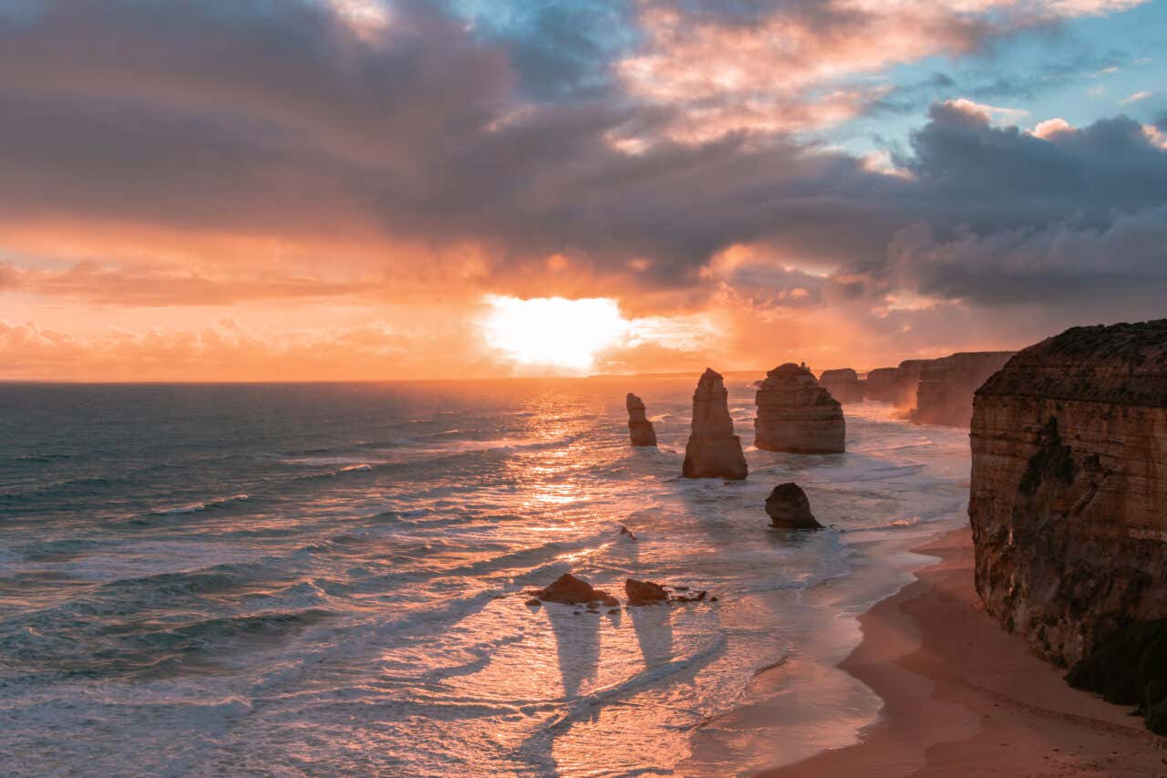 Sunset over the Twelve Apostles rock stacks along the Great Ocean Road in Victoria, Australia. The sun dips below a cloudy horizon, casting a bright orange glow over the ocean and illuminating the waves crashing against the beach and the towering limestone stacks.