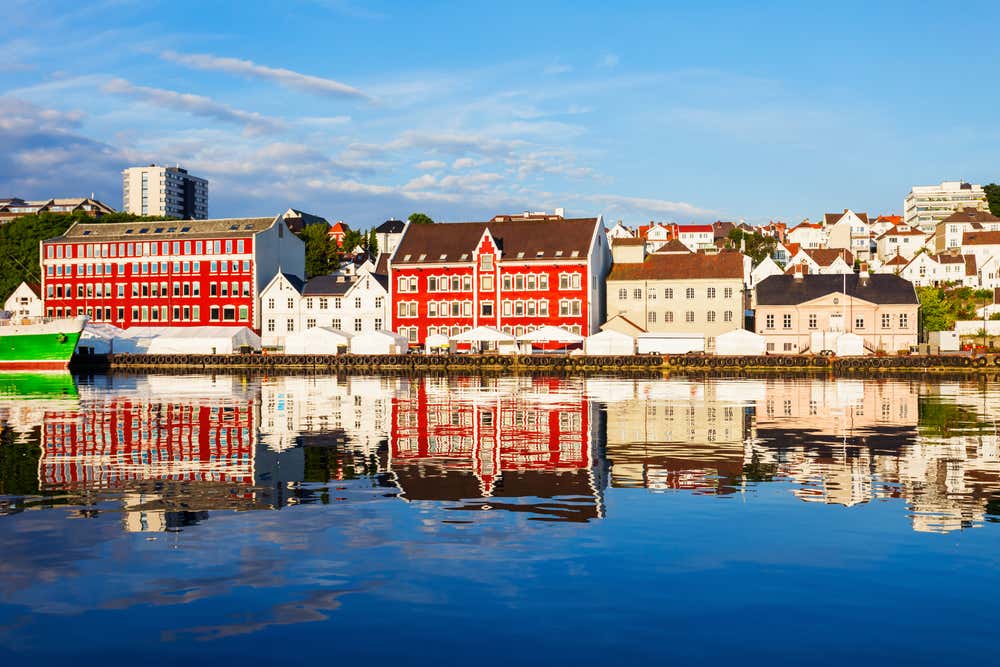 Vista del porto di Stavanger, in Norvegia: una fila di edifici storici dai vivaci colori rosso e bianco si specchia perfettamente nelle acque calme e azzurre del porto. A sinistra, un peschereccio verde è ormeggiato alla banchina. Sullo sfondo, le colline residenziali della città si stagliano sotto un cielo azzurro con qualche nuvola, restituendo un'immagine simmetrica e luminosa tipica delle città portuali norvegesi
