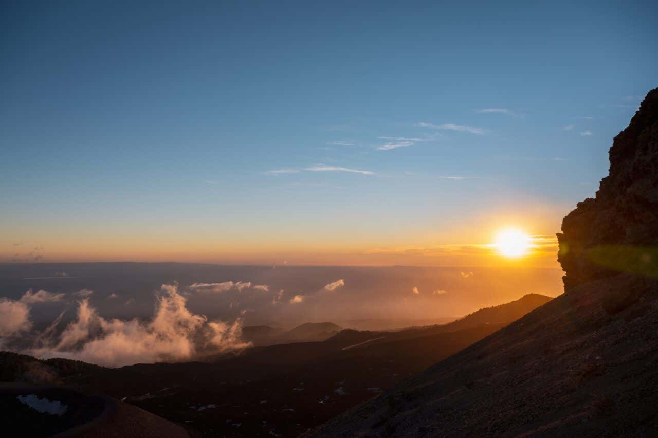 A sunrise or sunset viewed from a high altitude, likely on a mountain. The sun is a bright orb just above a hazy horizon, illuminating scattered clouds below. A rocky peak juts in from the right foreground, with a rugged landscape fading into the distance.