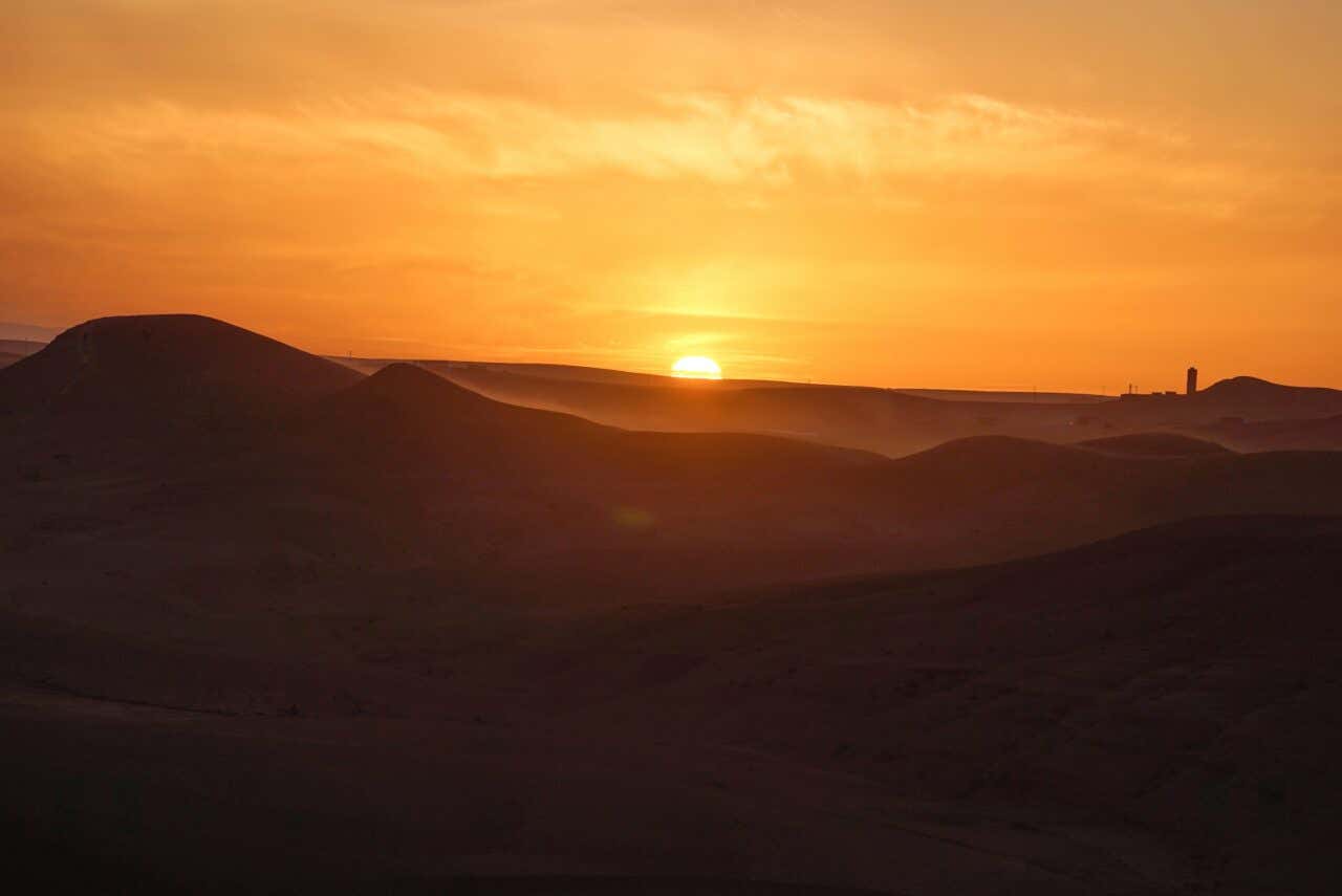 Sunset or sunrise over rolling sand dunes, possibly in a desert landscape. The sun is a bright, hazy orb just above the horizon, painting the sky a warm orange. The dark silhouettes of the dunes fill the foreground, with faint structures visible in the distance.