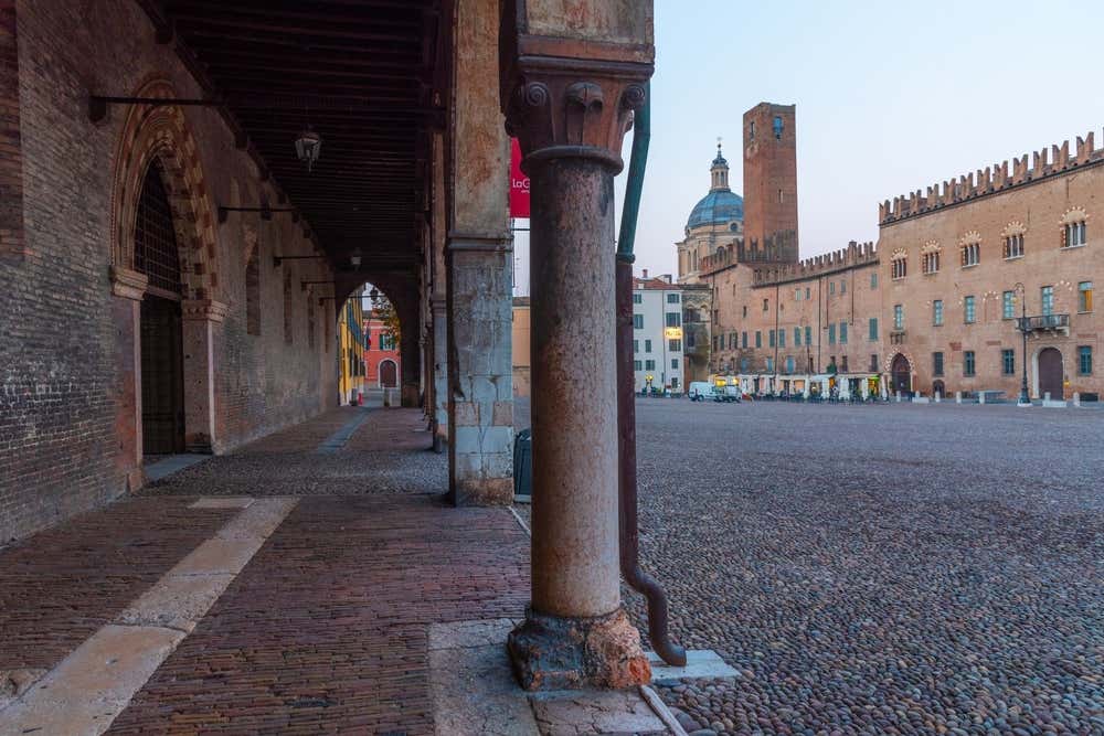 Piazza Sordello fotografata da sotto un arco medievale sorretto da colonne con capitelli decorati. La piazza è completamente pavimentata in ciottoli ed è circondata da palazzi medievali in mattoni, la luce sembra quella dell'alba, infatti la piazza è vuota