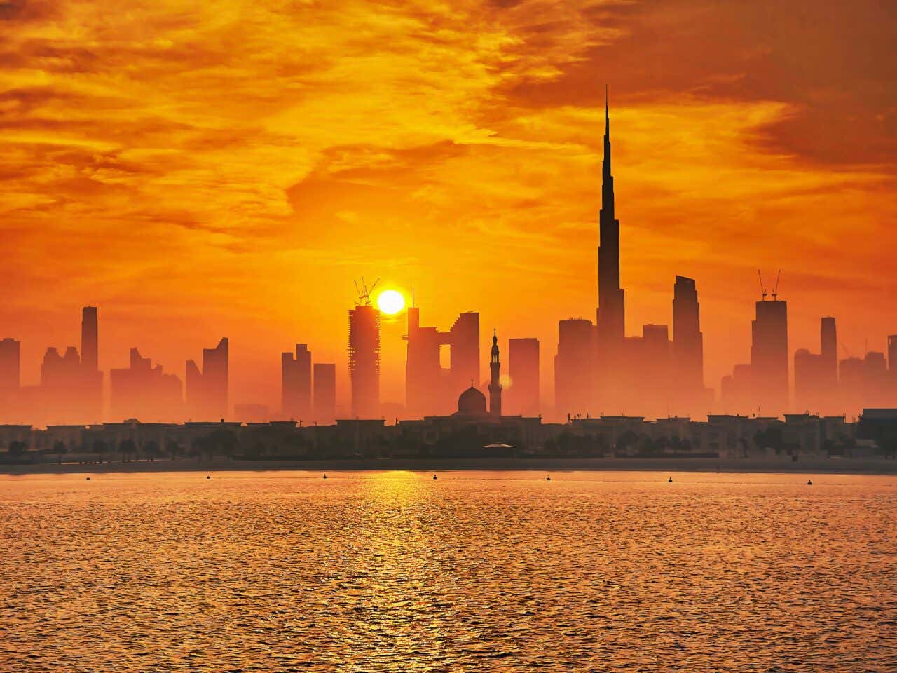 Sunset over the Dubai skyline, casting the city in silhouette against an intense orange and yellow sky. The Burj Khalifa is the tallest and most prominent structure, with other skyscrapers and a mosque dome visible, all reflected in the body of water in the foreground.