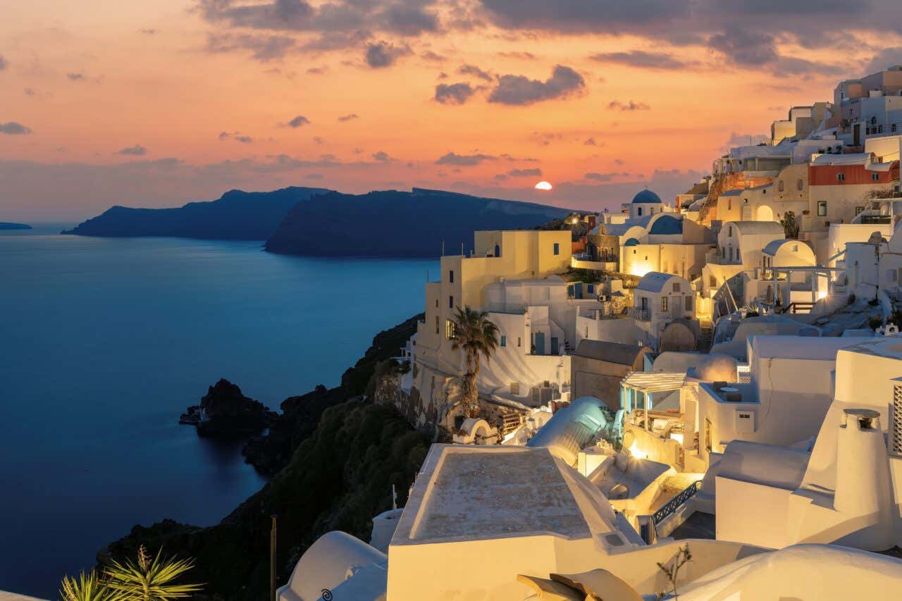 Sunset over Oia, Santorini, Greece. The iconic whitewashed buildings on the cliff face are illuminated by interior and street lights. The deep blue Aegean Sea and the caldera rim stretch out in the background under an orange and reddish twilight sky.