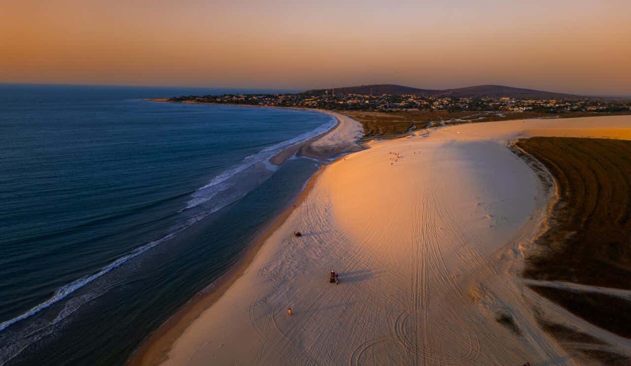 Sunset or sunrise over rolling sand dunes, possibly in a desert landscape. The sun is a bright, hazy orb just above the horizon, painting the sky a warm orange. The dark silhouettes of the dunes fill the foreground, with faint structures visible in the distance.