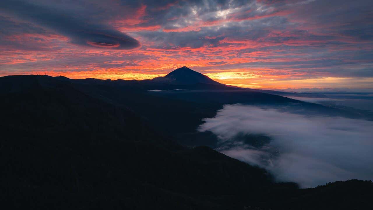A dramatic sunrise or sunset over Mount Teide silhouetted against a vibrant orange and red horizon, while clouds and mist cling to the dark, forested slopes in the foreground.