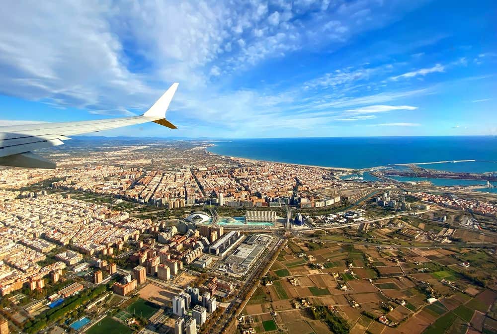 Veduta aerea di Valencia, Spagna, ripresa dal finestrino di un aereo in fase di atterraggio o decollo. L'ala dell'aeromobile incornicia il paesaggio urbano della città, con il suo tessuto edilizio denso e ordinato che si estende fino alla costa mediterranea. In primo piano spicca la Città delle Arti e delle Scienze con le sue iconiche strutture architettoniche futuristiche. Sullo sfondo, il porto di Valencia e il Mar Mediterraneo brillano sotto un cielo azzurro con nuvole sparse