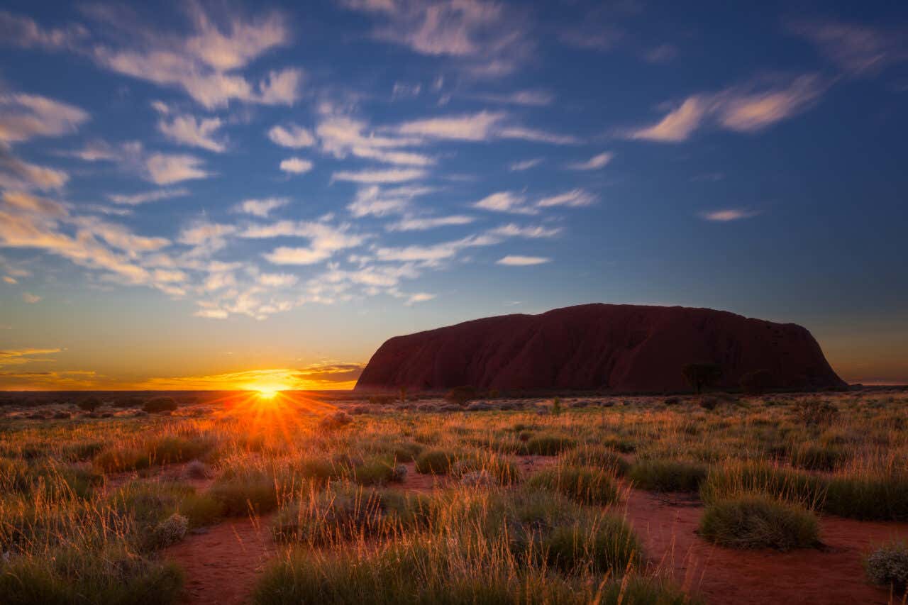 Sunrise over Uluru (Ayers Rock) in the Australian outback. The massive sandstone monolith is silhouetted against a brilliant sky, while the rising sun casts a golden glow on the dry, grassy foreground and red dirt path.