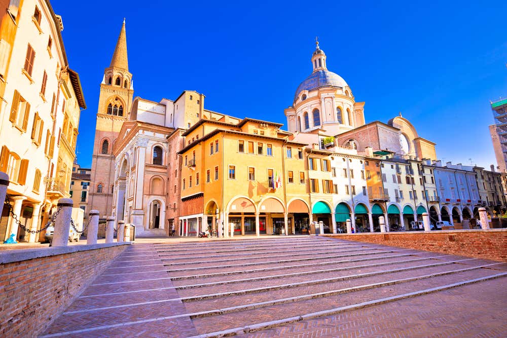 Piazza delle Erbe a Mantova con edifici storici colorati, portici ad archi, la Basilica di Sant'Andrea con la sua cupola e cielo azzurro
