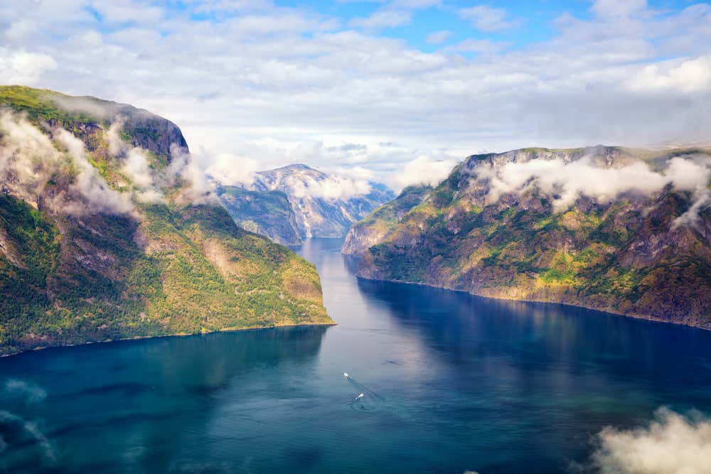 Vista aerea del Sognefjord, il fiordo più lungo della Norvegia: acque blu profondo si estendono tra ripide pareti rocciose coperte di vegetazione verde, avvolte da nuvole basse. In lontananza il fiordo si restringe verso l'orizzonte, mentre una piccola imbarcazione solca le acque silenziose sotto un cielo parzialmente nuvoloso