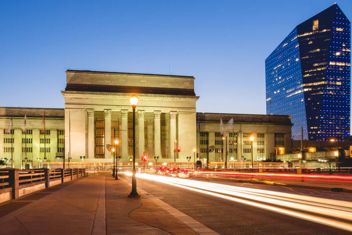 Exteriores de un edificio de fachada clásica con grandes columnatas iluminadas por la noche y junto a un rascacielos