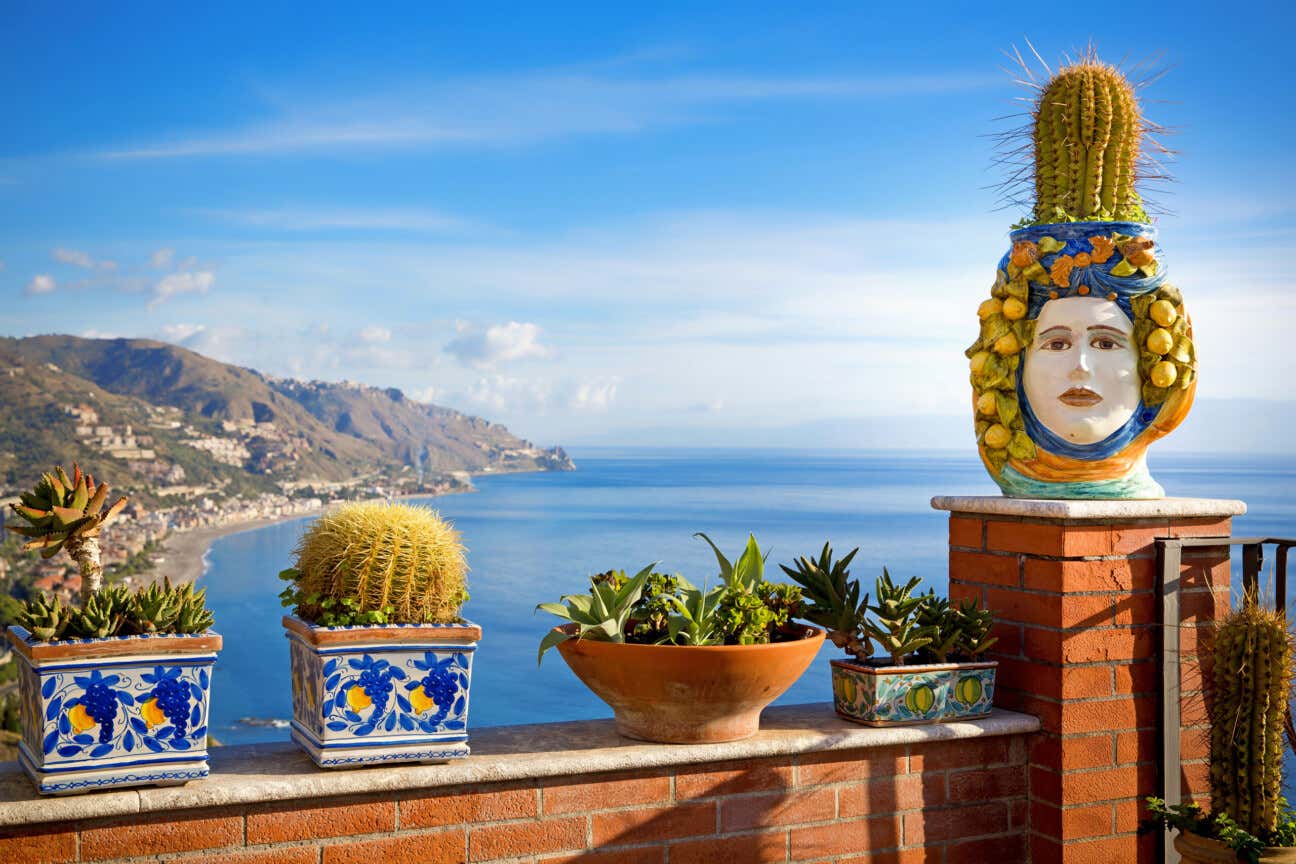 Vista sul golfo di Taormina con il mare cristallino e il cielo blu, mentre in primo piano vasi decorativi in ceramica tipici della Sicilia
