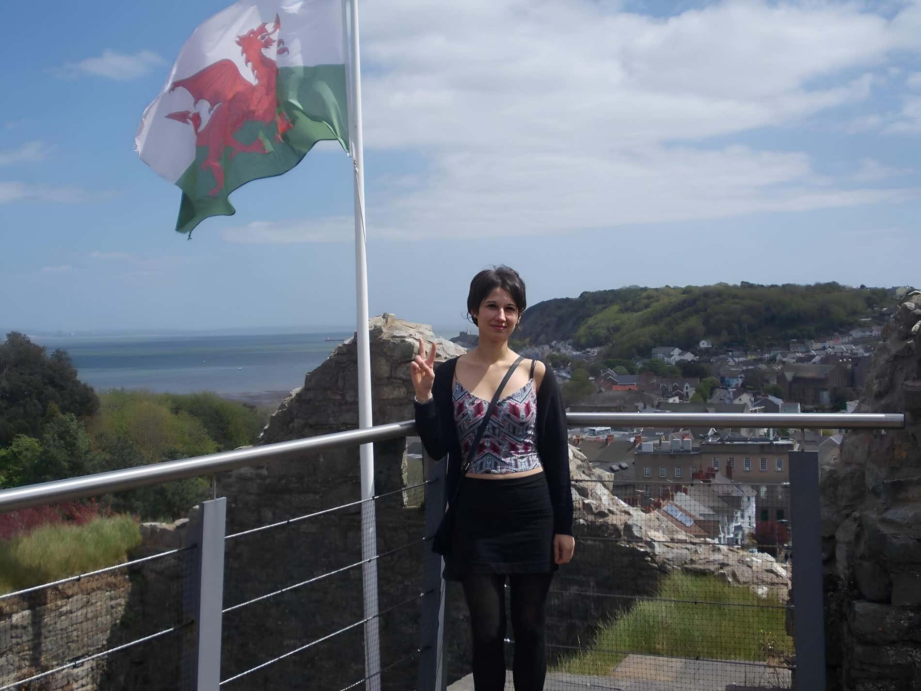 Una ragazza sorride sul belvedere di un castello, con il paesaggio e una bandiera del Galles alle spalle