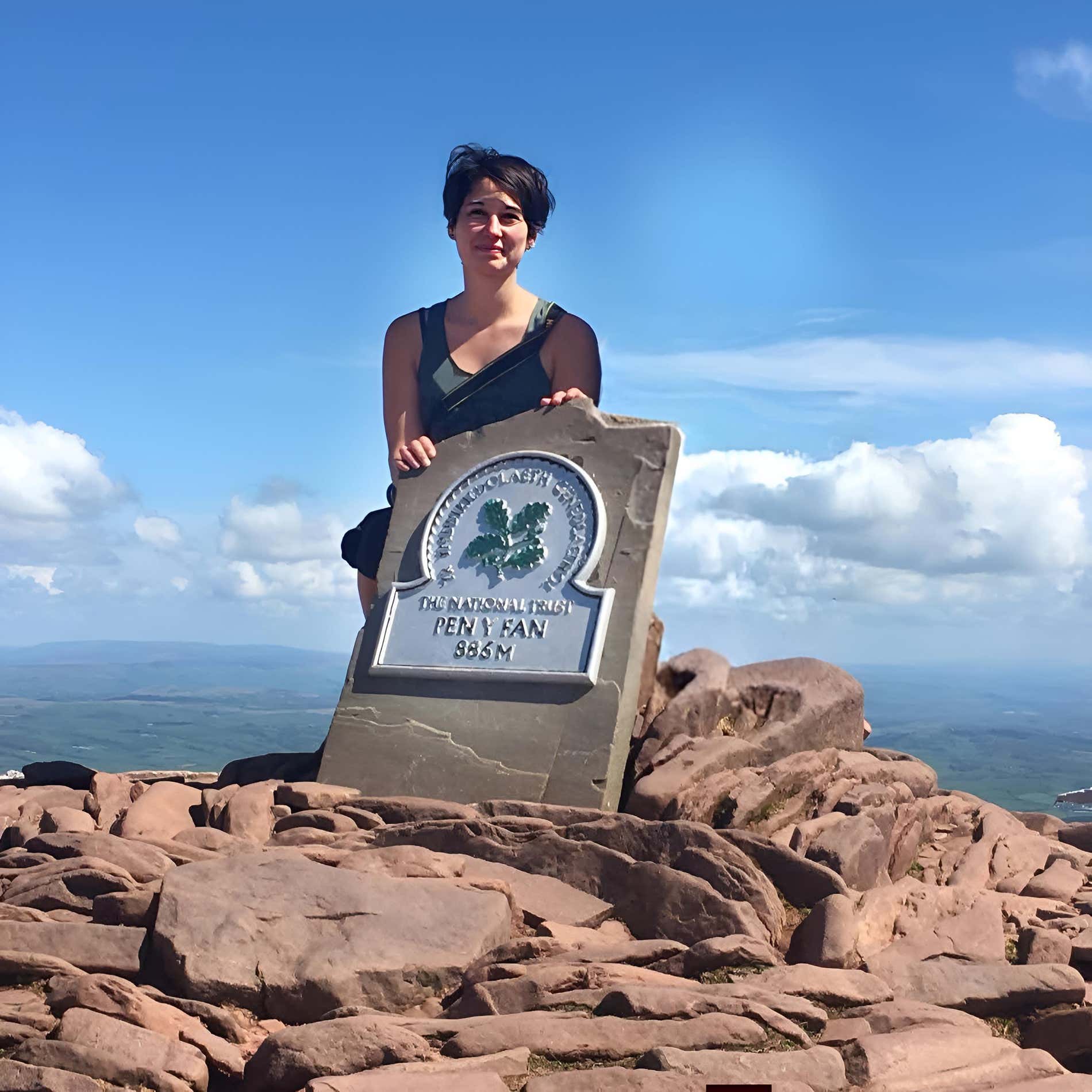 Una donna sorride sulla cima del Pen-y-Fan, accanto a un cartello che ne indica l'altezza