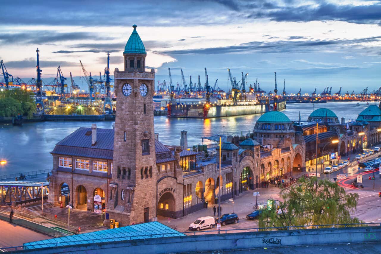 The illuminated clock tower and green-domed buildings of the St. Pauli Piers in Hamburg overlook a bustling harbor filled with cranes and ships under a dramatic evening sky.