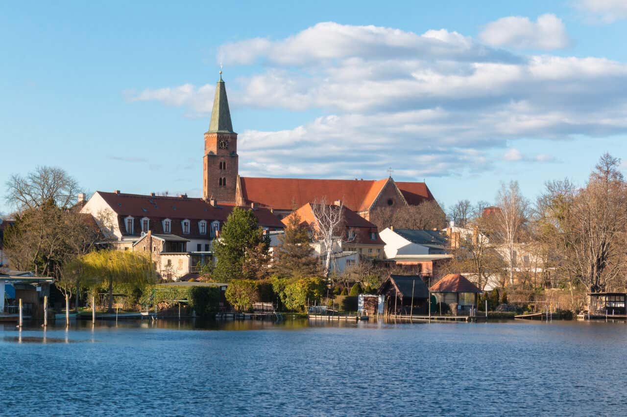 A view from across the Havel river shows the red-roofed buildings and pointed green spire of the island church in Werder, nestled among lush trees and waterfront gardens.