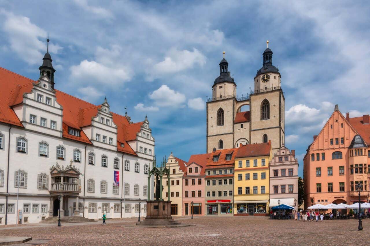 The historic Market Square in Lutherstadt Wittenberg, showing the ornate white Town Hall on the left and the towering twin spires of the Stadtkirche (City Church) rising behind a row of colorful, multi-story buildings under a cloudy blue sky.